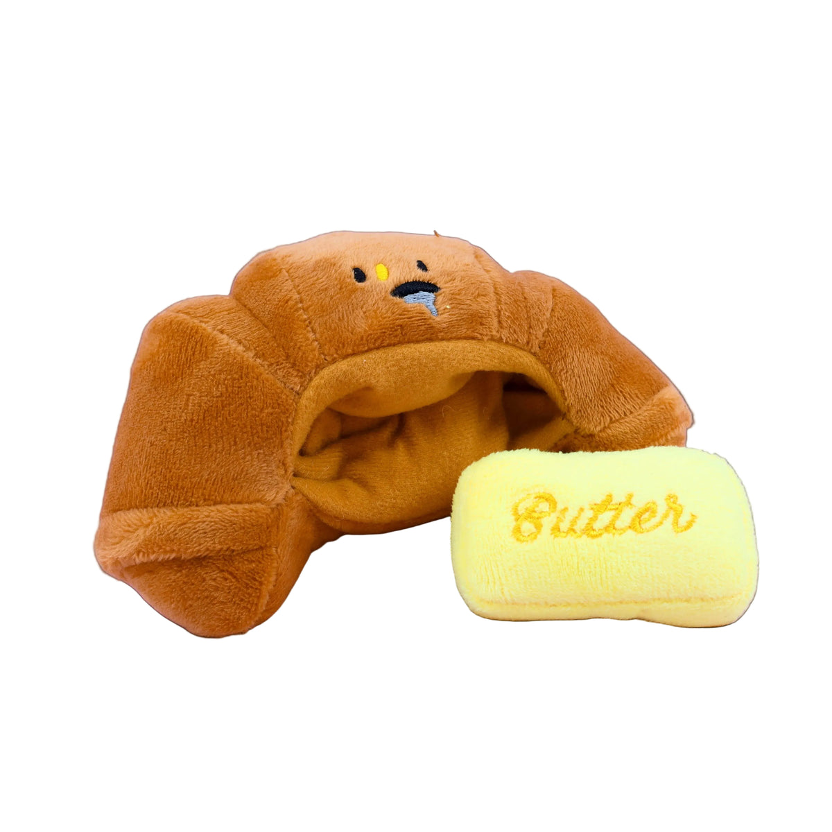 Plush toy resembling a brown croissant with a small yellow rectangular pillow labeled 'Butter' on a black background.